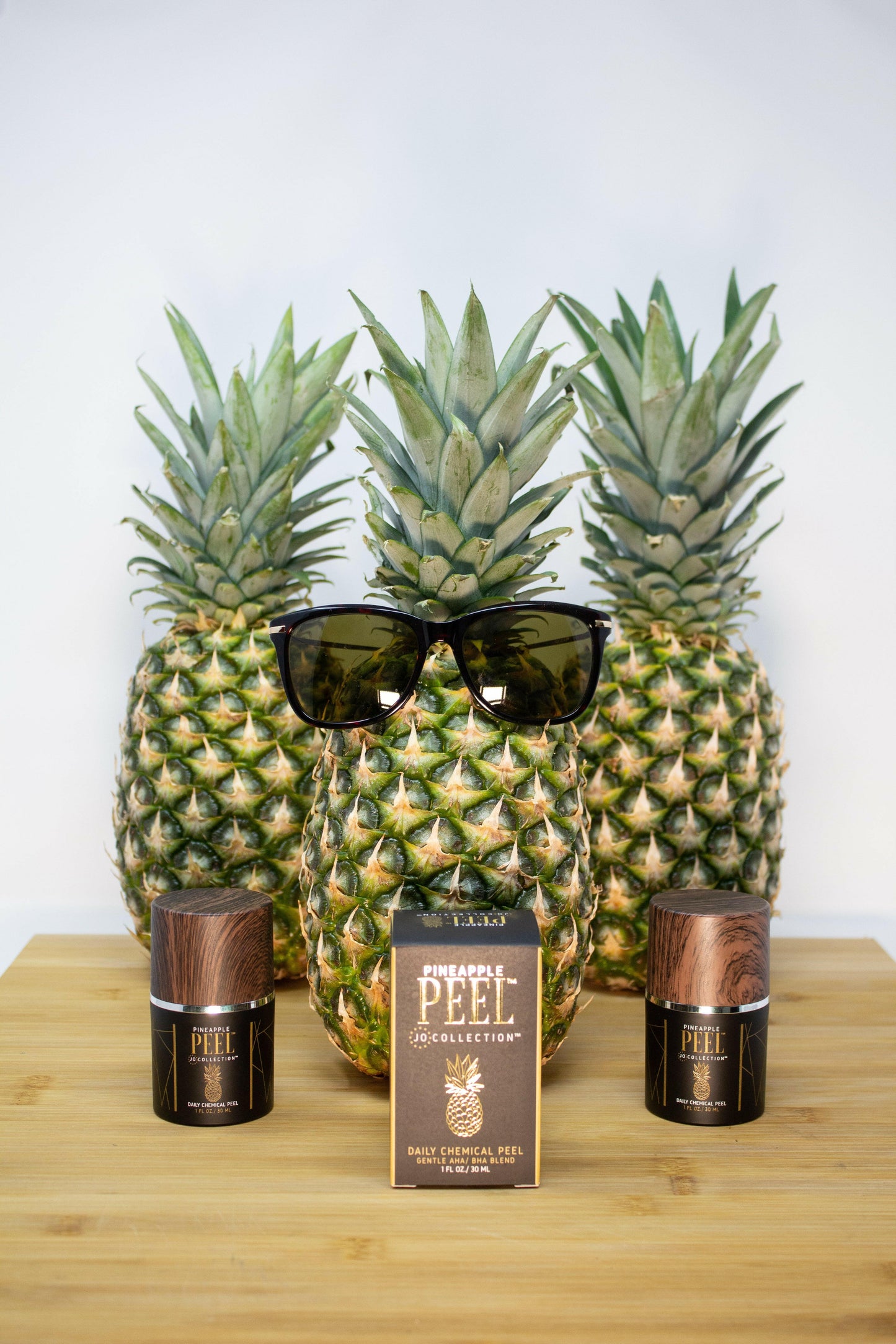 Three pineapples with sunglasses and two bottles on a wooden surface
